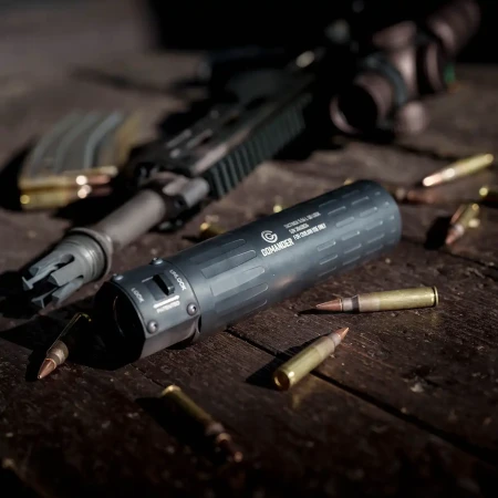 Tłumik Gomander TACTINOX QD-LOCK L 5.56 mm na stole obok karabinka i amunicji kalibru 5.56.